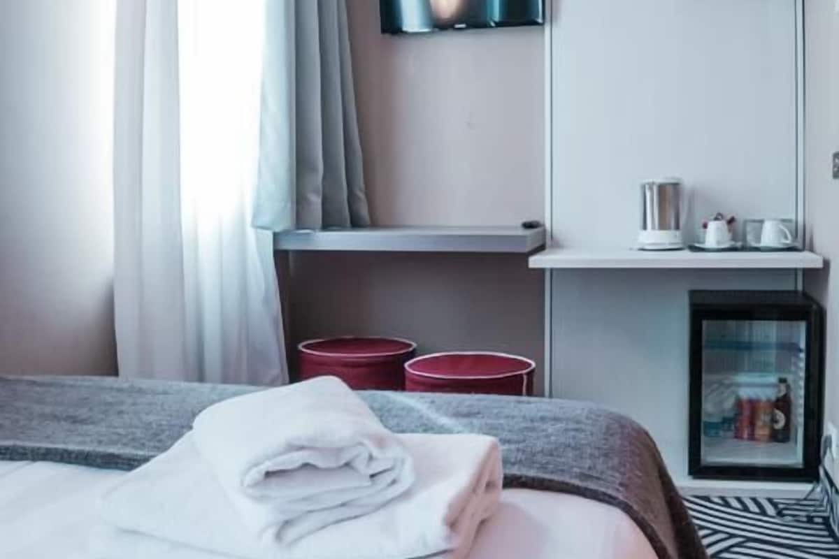 A hotel room with a bed, a TV mounted on the wall, a small table with a kettle, and a red trash bin.