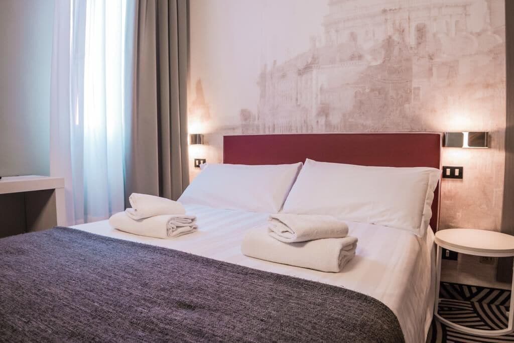 Una cama bien hecha con ropa de cama y toallas blancas, un cabecero rojo y una lámpara fijada a la pared.