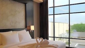 A hotel room with a large bed, a nightstand, a lamp, and a view through a large window. - Grand Karlita Purwokerto (Purwokerto)