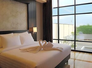 A hotel room with a large bed, a nightstand, a lamp, and a view through a large window. - Grand Karlita Purwokerto (Purwokerto)
