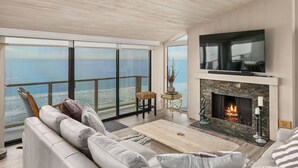A spacious living room with a large window overlooking the sea, a fireplace, and a flat-screen TV mounted on the wall. - Sea For Miles Top Floor Oceanfront Rental - A/C, Beach Access, Pool (Solana Beach)