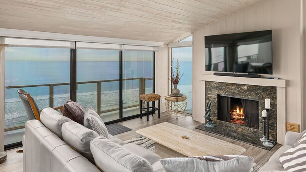 A spacious living room with a large window overlooking the sea, a fireplace, and a flat-screen TV mounted on the wall. - Sea For Miles Top Floor Oceanfront Rental - A/C, Beach Access, Pool (Solana Beach)