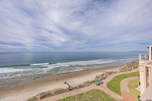 Vista a la playa desde un balcón, con un paseo pavimentado, una zona verde y un edificio con techo inclinado.