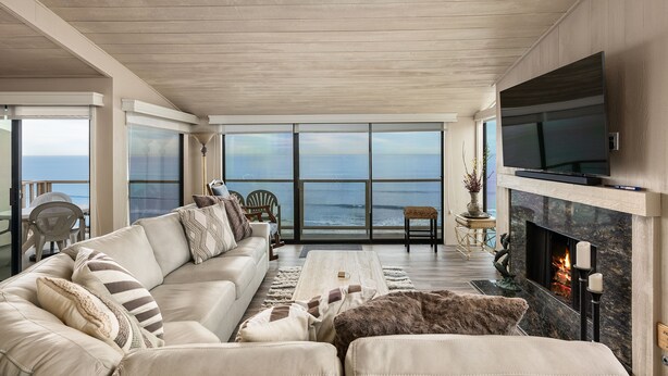 A spacious living room with a large sofa, a flat-screen TV, and a fireplace. - Sea For Miles Top Floor Oceanfront Rental - A/C, Beach Access, Pool (Solana Beach)