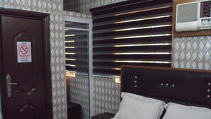 Habitación de hotel con cama, puerta con cartel de 'No Fumar', ventana con persianas y aire acondicionado montado en la pared.