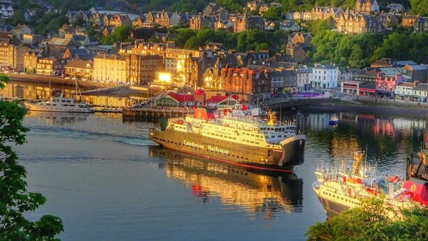 Un gran crucero llamado Caledonian MacBrayne atracado en un puerto, con un paisaje urbano al fondo.