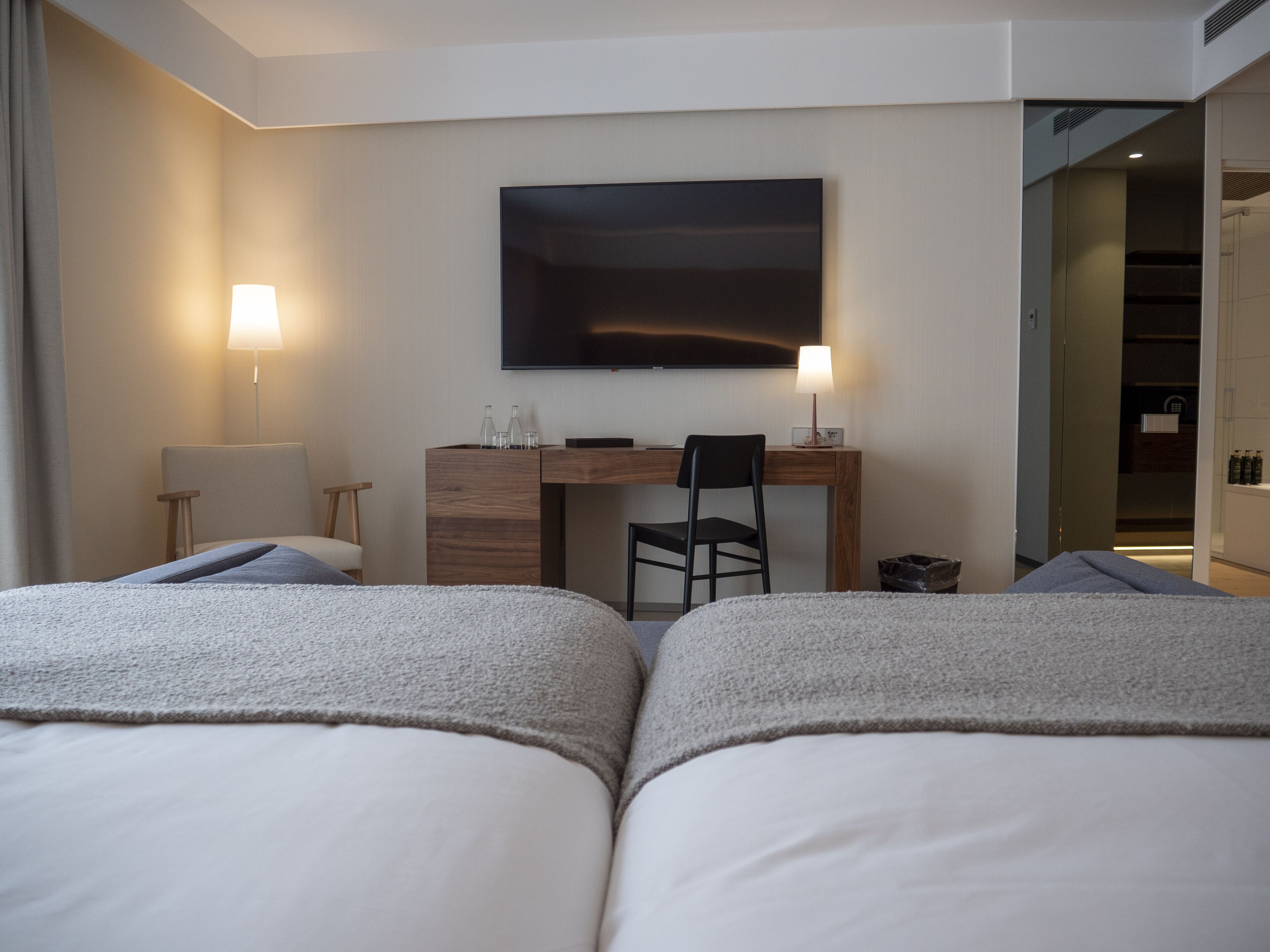 A hotel room with two beds, a desk, a chair, a TV, and a lamp.