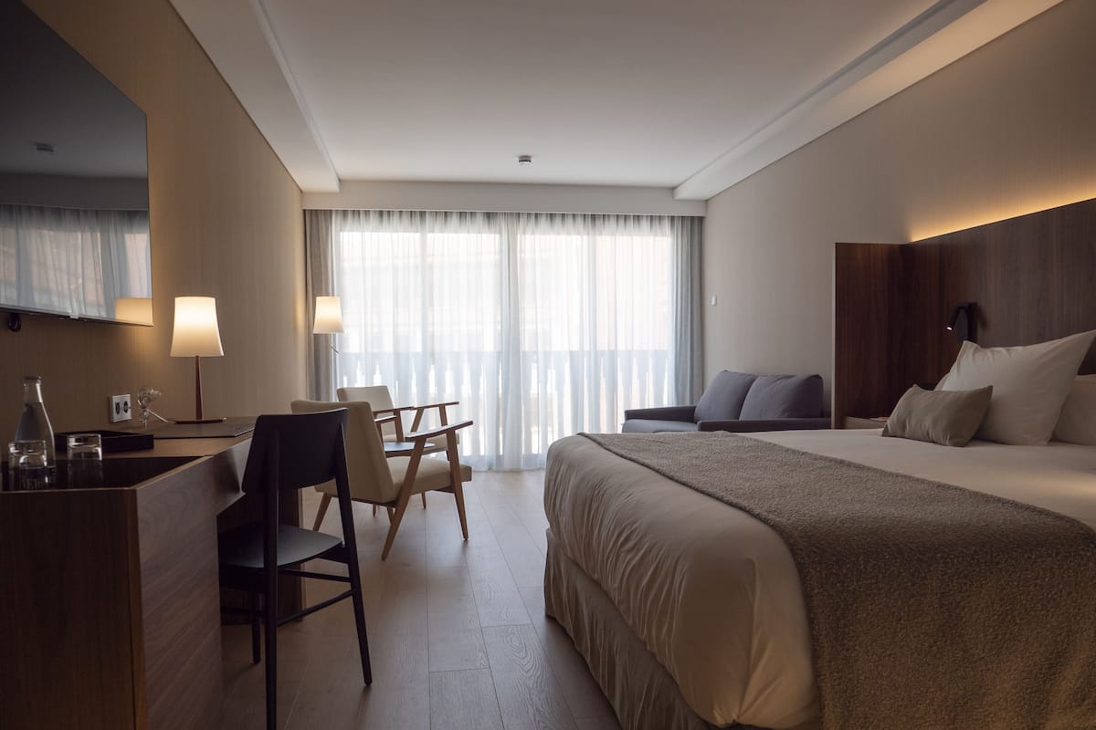 Una habitación de hotel con cama, escritorio, silla, lámpara y una ventana grande con cortinas.
