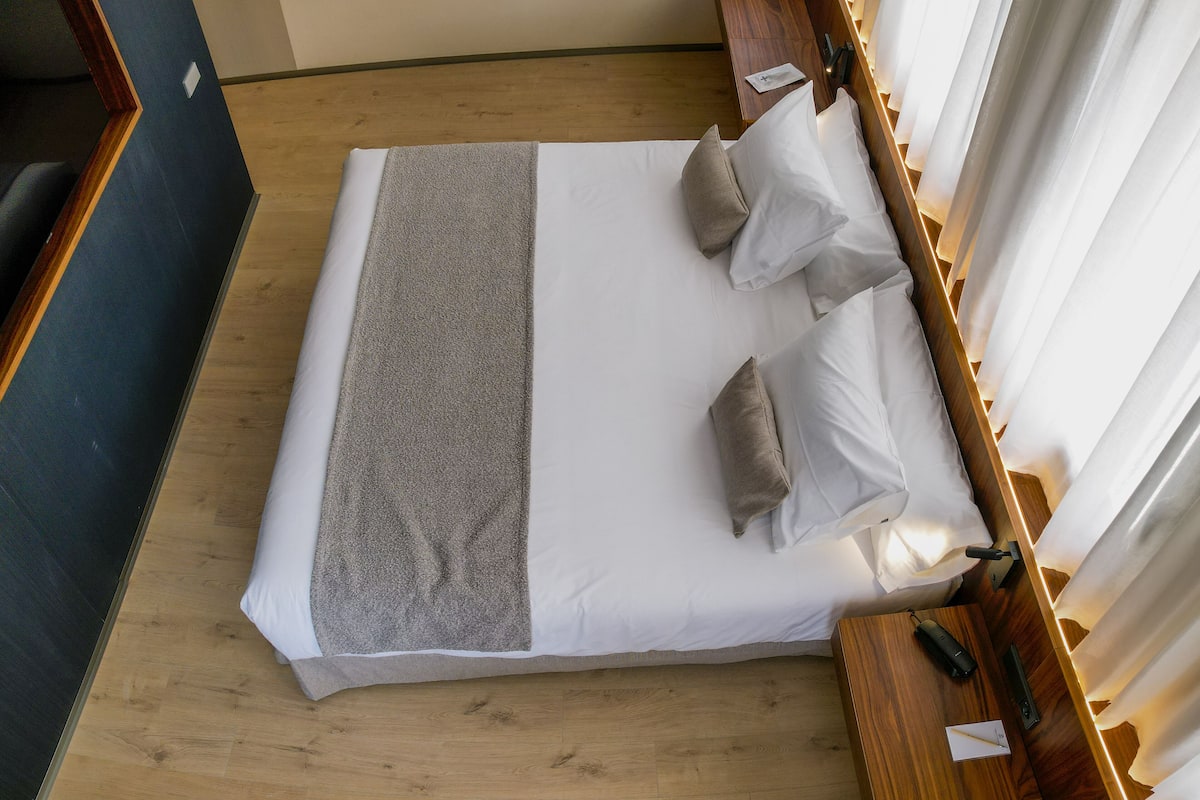 Una cama bien hecha con una manta gris y dos almohadas, una mesita de noche de madera con una lámpara, y una ventana con cortinas blancas.