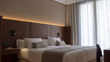 A modern hotel room with a large bed, wooden paneling, a desk, and a view of the city through sheer curtains.