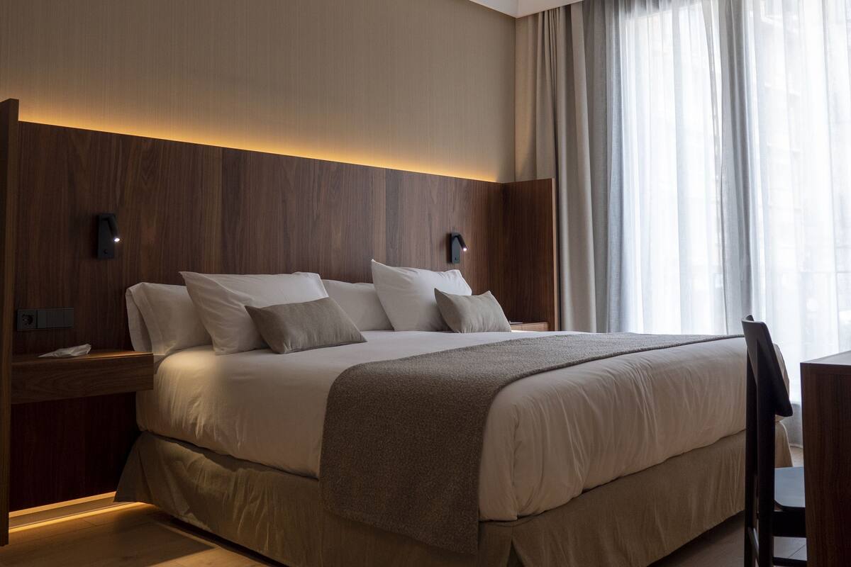 Un dormitorio moderno con una cama grande, cabecera de madera, mesitas de noche y vistas a la ciudad a través de cortinas traslúcidas.