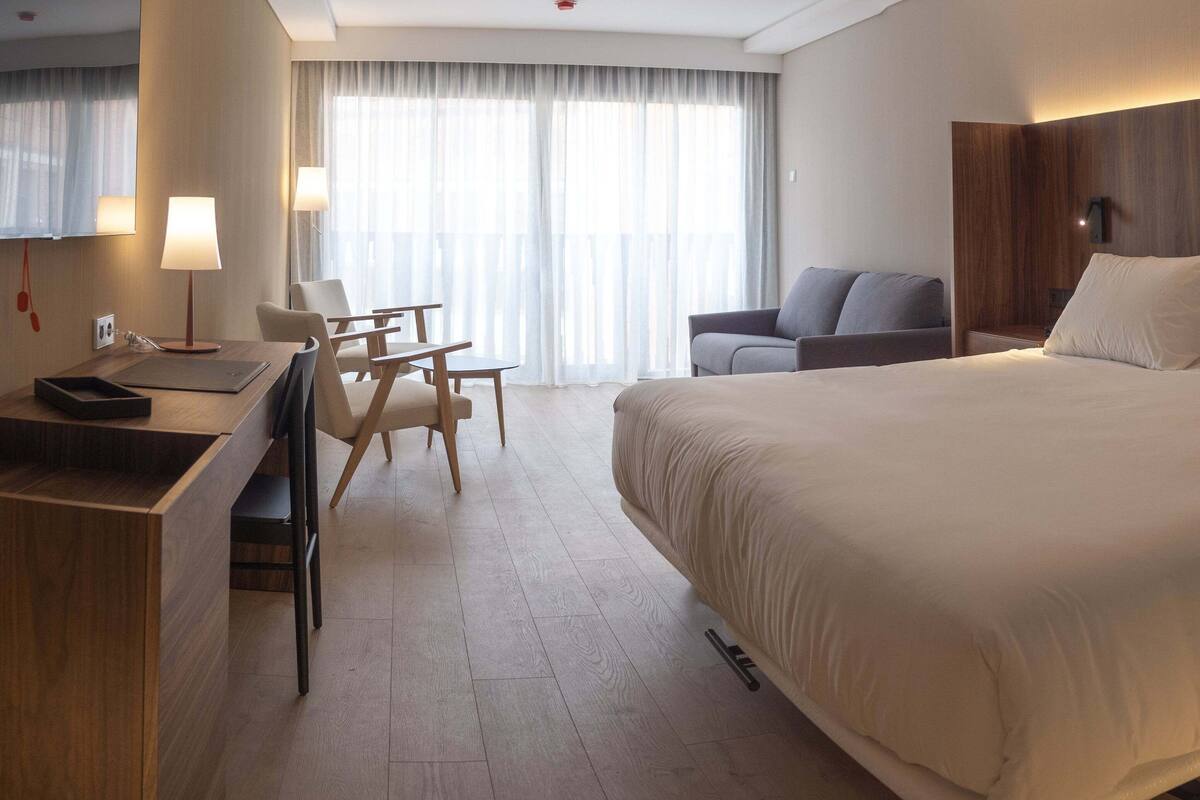 Habitación de hotel con una cama grande, un escritorio con lámpara, una silla y vistas al exterior a través de amplias ventanas.