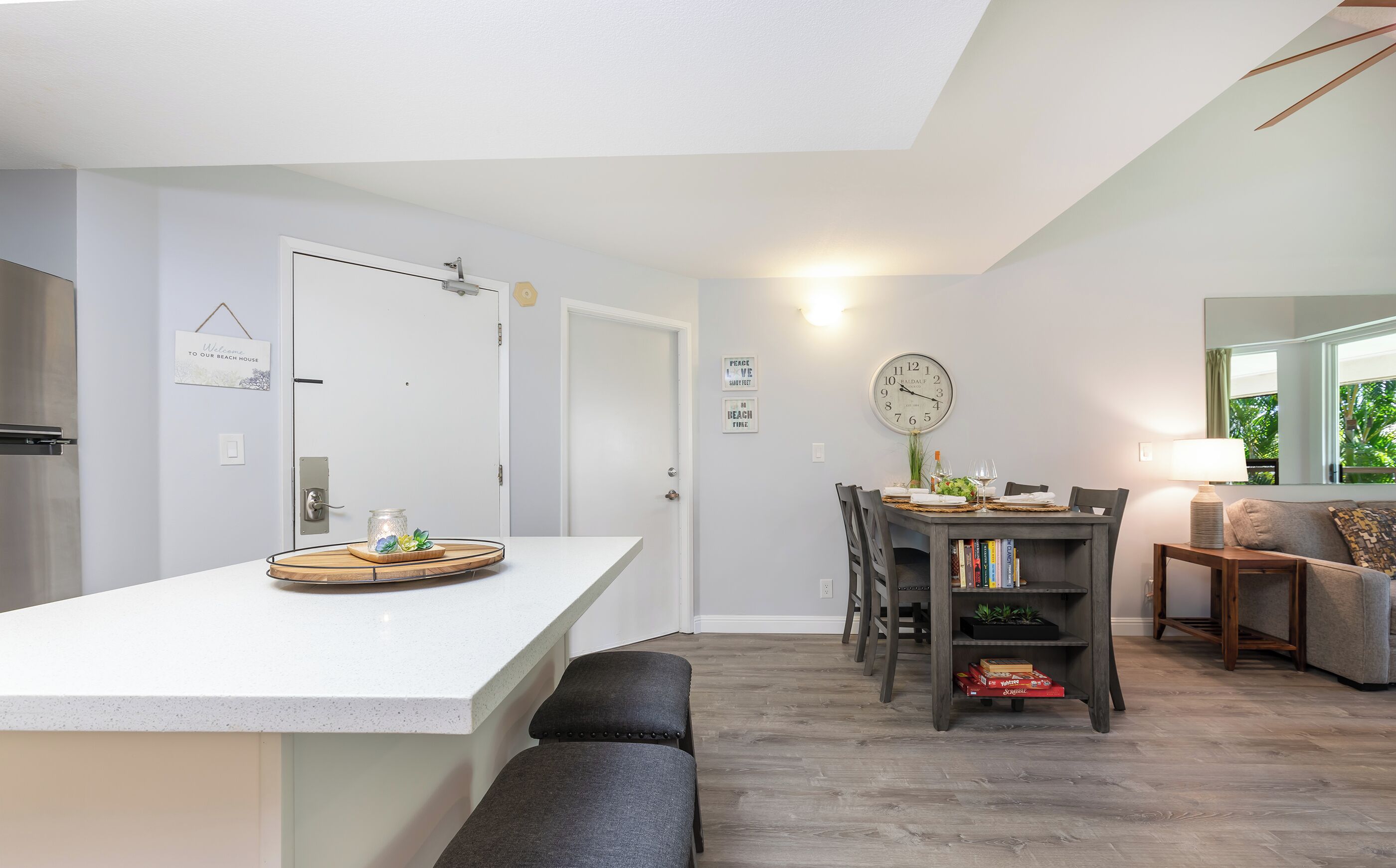 A modern kitchen with a white countertop, a dining area with a wooden table and chairs, a clock on the wall, and a cozy seating area with a sofa and a lamp.