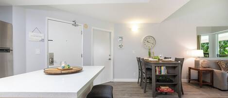 A modern kitchen with a white countertop, a dining area with a wooden table and chairs, a clock on the wall, and a cozy seating area with a sofa and a lamp.