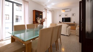 A modern living room with a glass dining table, beige chairs, a sofa set, a flat-screen TV, and a wooden cabinet. - Large 3bedroom apartment near rocky beach MCRE1-1 (Sliema)
