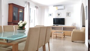 A modern living room with a glass-top dining table, beige sofas, a wooden cabinet, a flat-screen TV, and framed pictures on the wall. - Large 3bedroom apartment near rocky beach MCRE1-1 (Sliema)