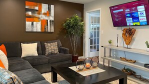 Un salón moderno con un sofá oscuro, una mesa de centro y un televisor de pantalla plana grande montado en la pared que muestra la interfaz de un servicio de streaming.