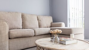 En stue med en beige sofa, et rundt træbord, en vase med visne blomster, et stearinlys og en bog.