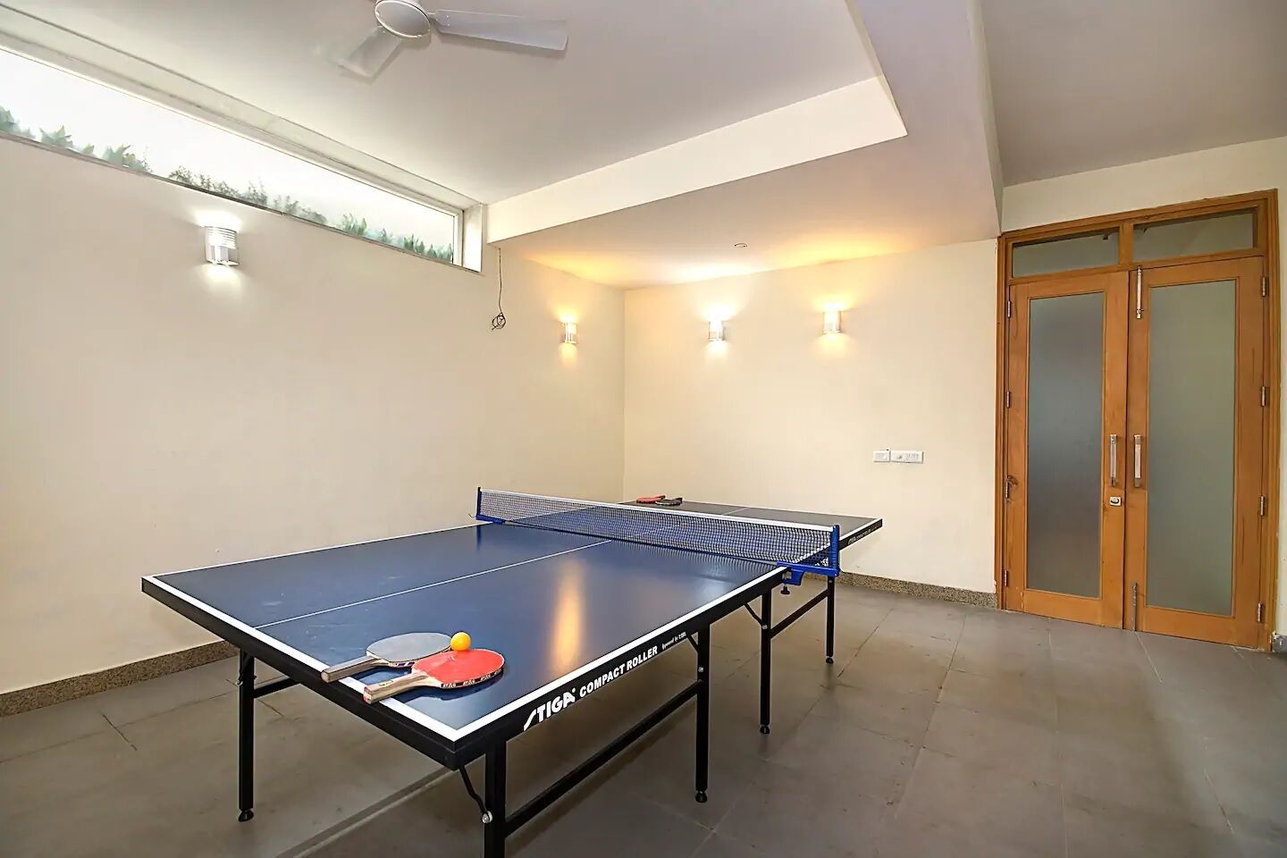A ping pong table with two paddles and a ball, a ceiling fan, and a door with frosted glass panels.