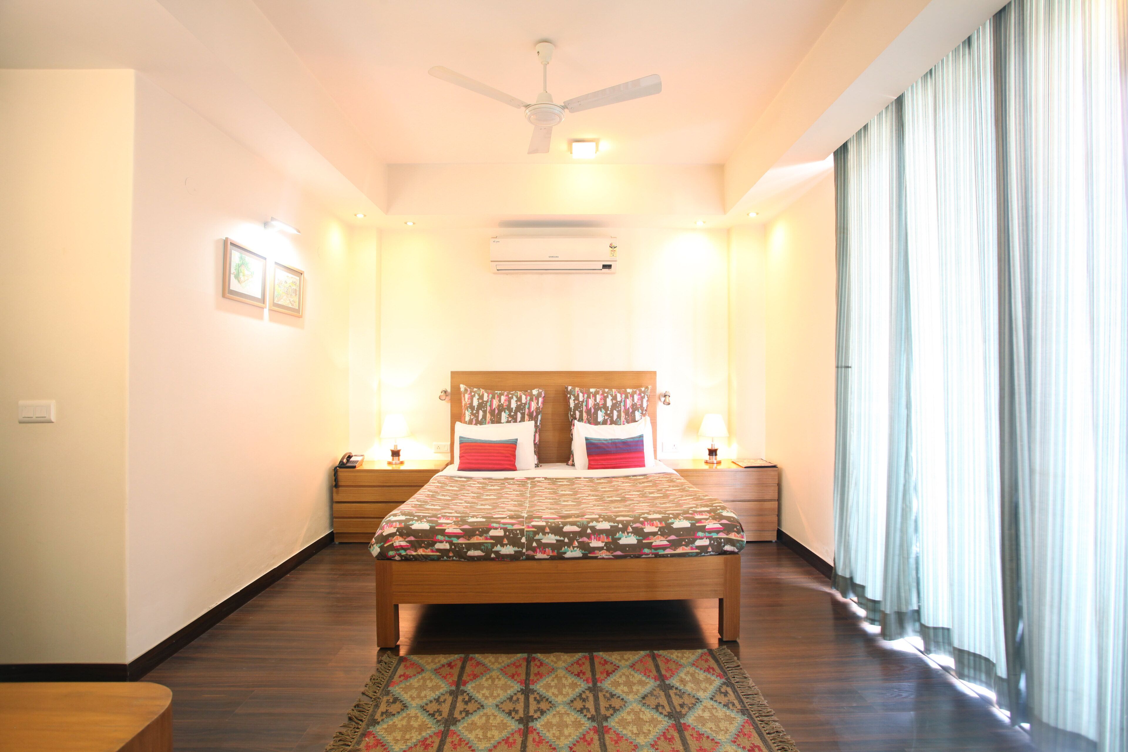 A hotel room with a bed, bedside tables, a ceiling fan, an air conditioner, and a window with curtains.