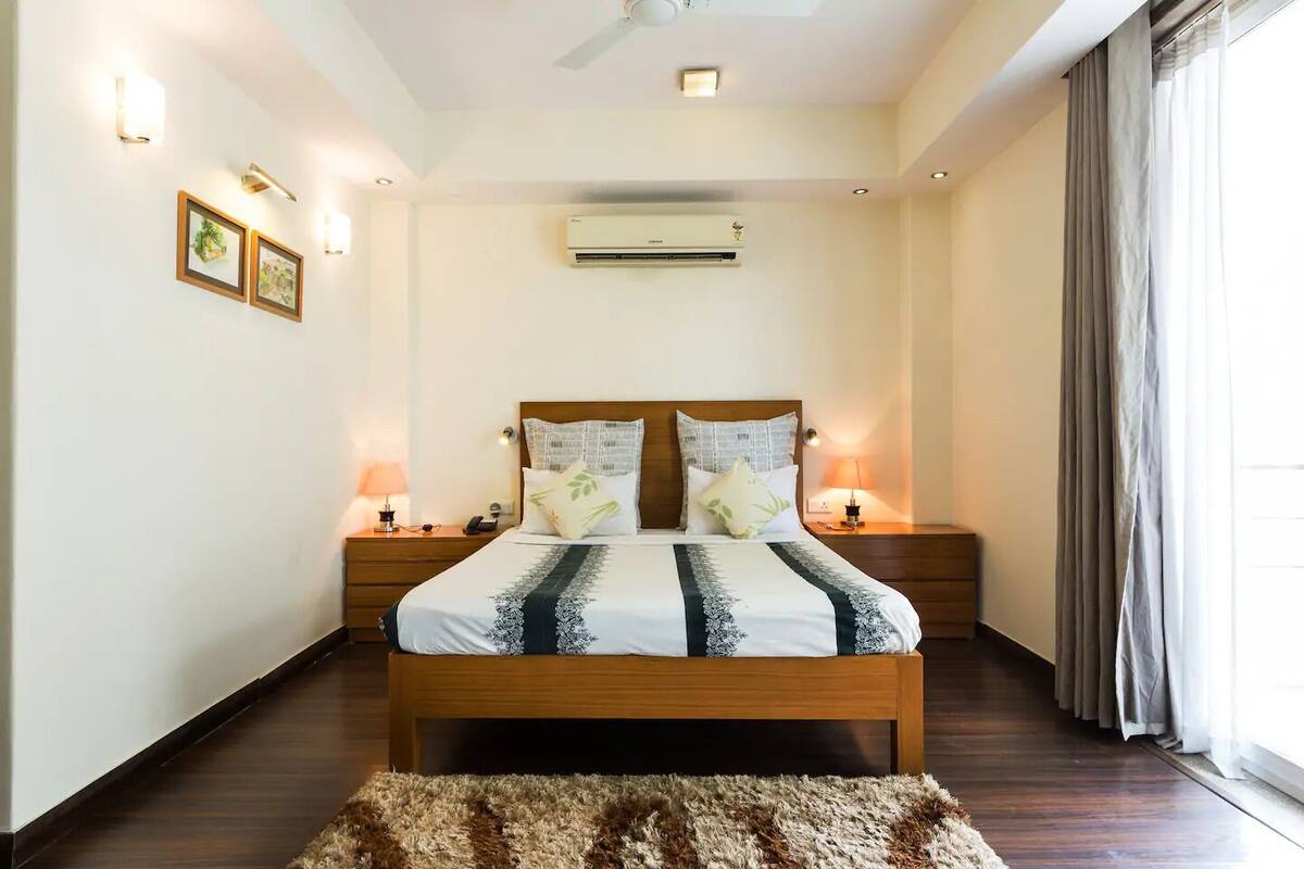 Un dormitorio con cama de madera, dos mesitas de noche con lámparas, un equipo de aire acondicionado y vistas al exterior a través de una ventana con cortinas.