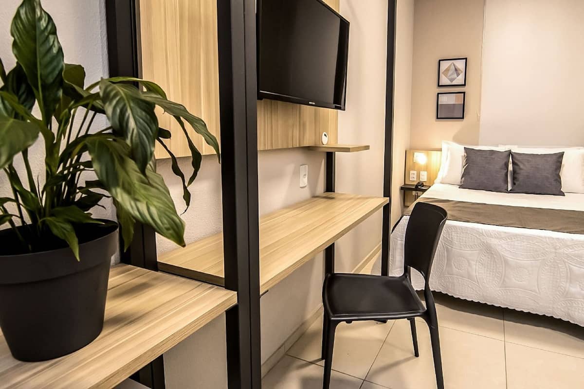 A hotel room with a bed, a desk, a chair, a TV, and a potted plant.