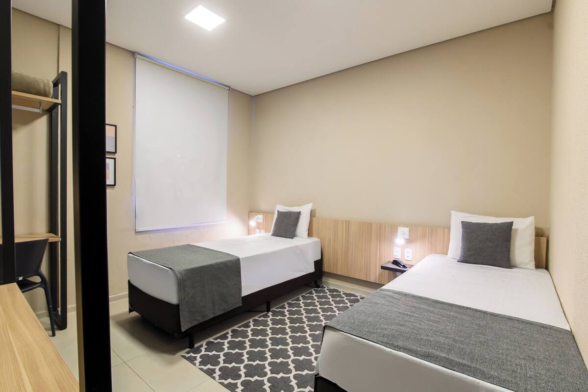 A hotel room with two beds, a desk, a chair, a projector screen, and a patterned rug.