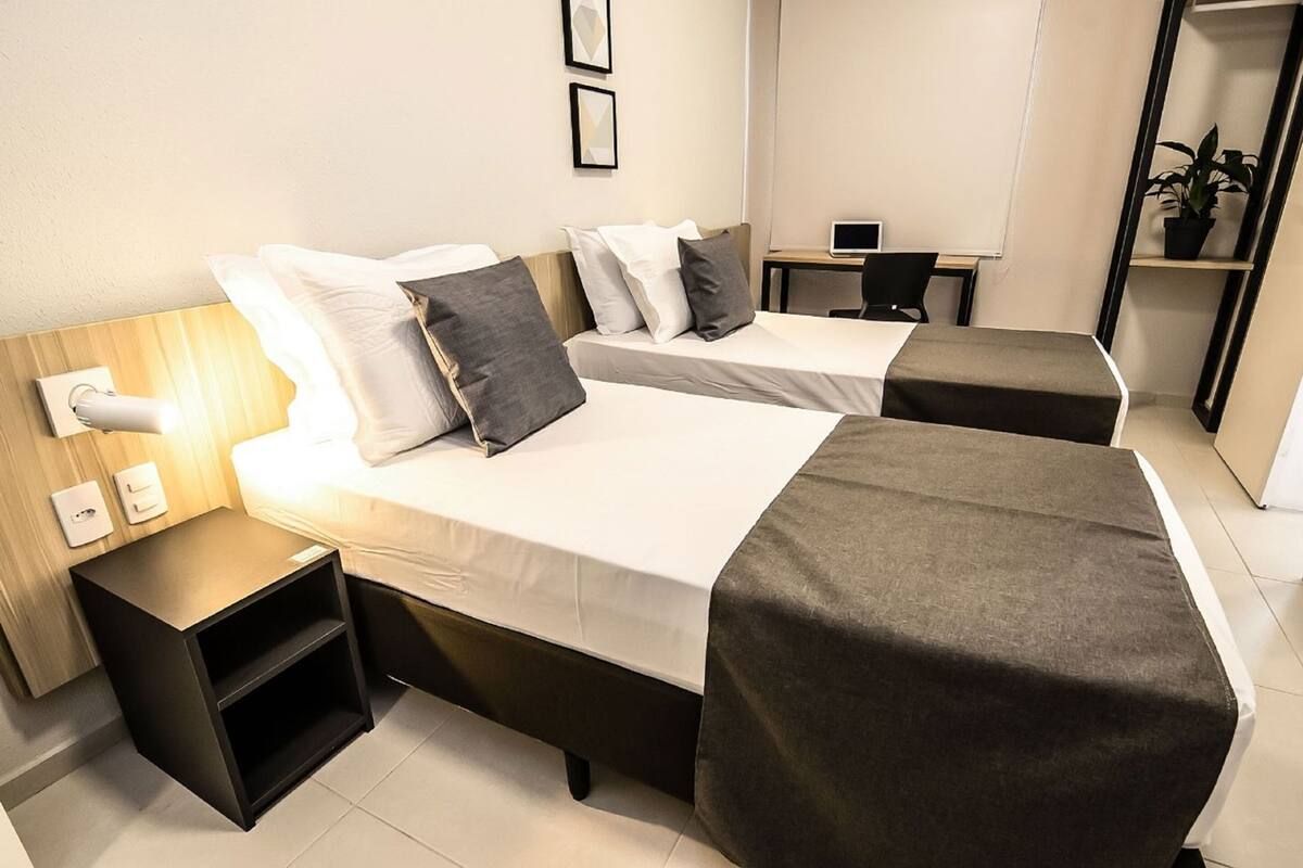 A hotel room with two beds, a desk, a chair, a lamp, and a potted plant.