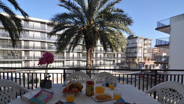 Une table sur le balcon, avec des éléments pour le petit-déjeuner, un livre et une vue sur les bâtiments et les palmiers.