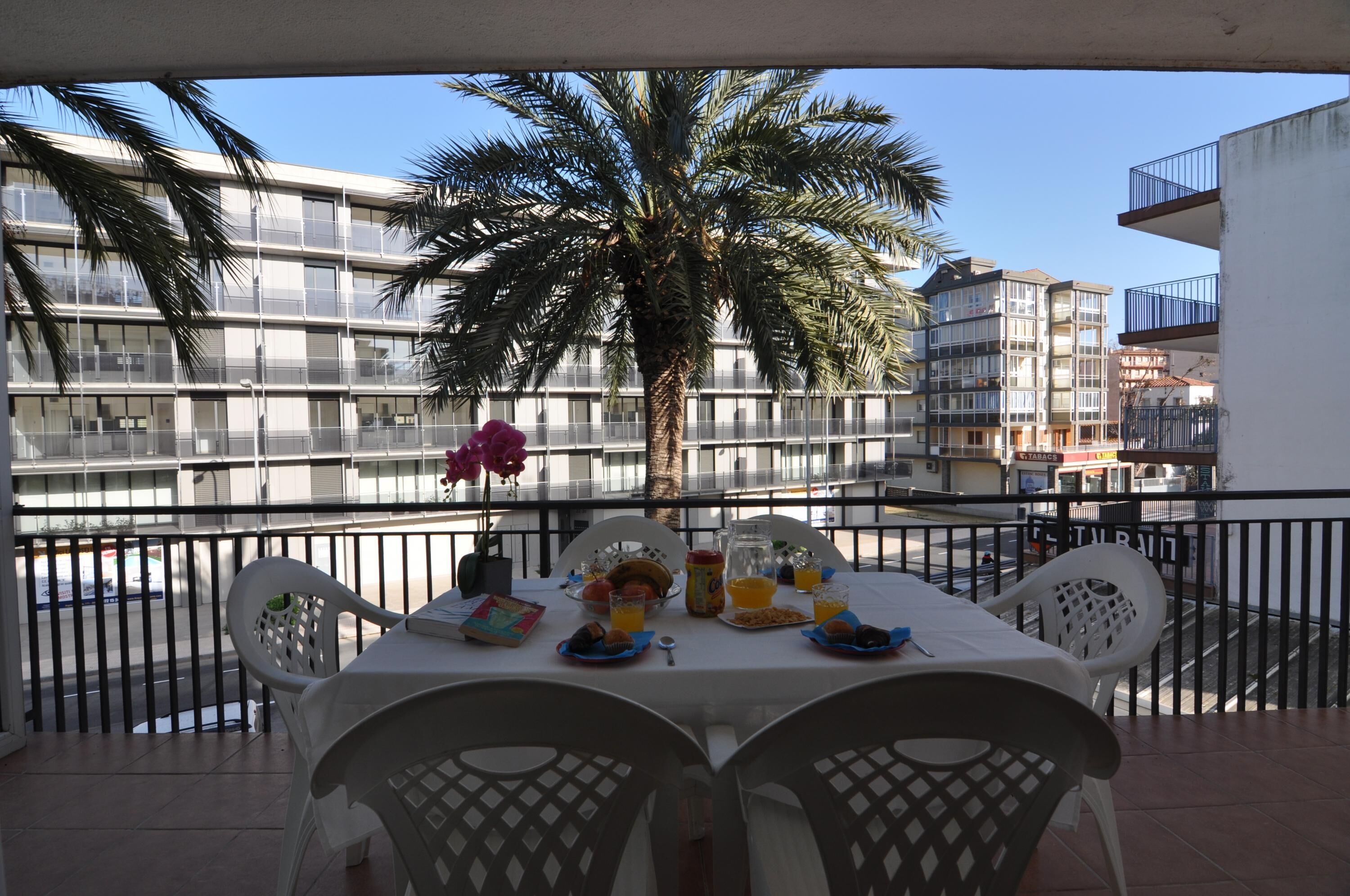 A balcony with a table set for breakfast, overlooking a palm tree and modern buildings.