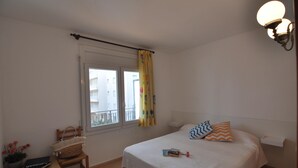 Une chambre à coucher comprenant un lit, une table de chevet, une fenêtre avec des rideaux, une chaise, une petite table avec un panier et des livres, et un luminaire fixé au mur.