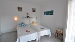 A small, clean room with two single beds, a small table, and a chair. There are wall-mounted lights and a shelf with a vase of flowers. - Welcome to our cozy apartment for rent in Roses, Costa Brava! Just 300 meters from the beautiful Santa Margarita beach. (Roses)