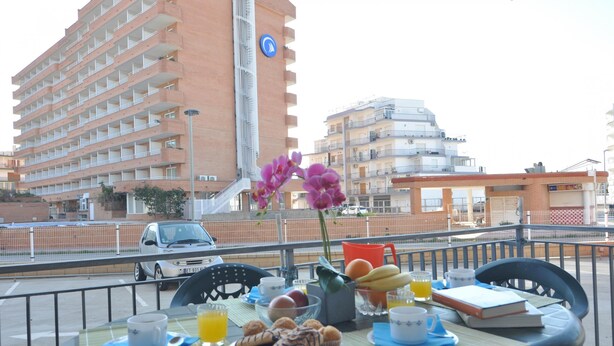 A balcony table with breakfast items, a flower vase, and a view of a tall building and a car in the background. - Welcome to our cozy apartment for rent in Roses, Costa Brava! Just 300 meters from the beautiful Santa Margarita beach. (Roses)