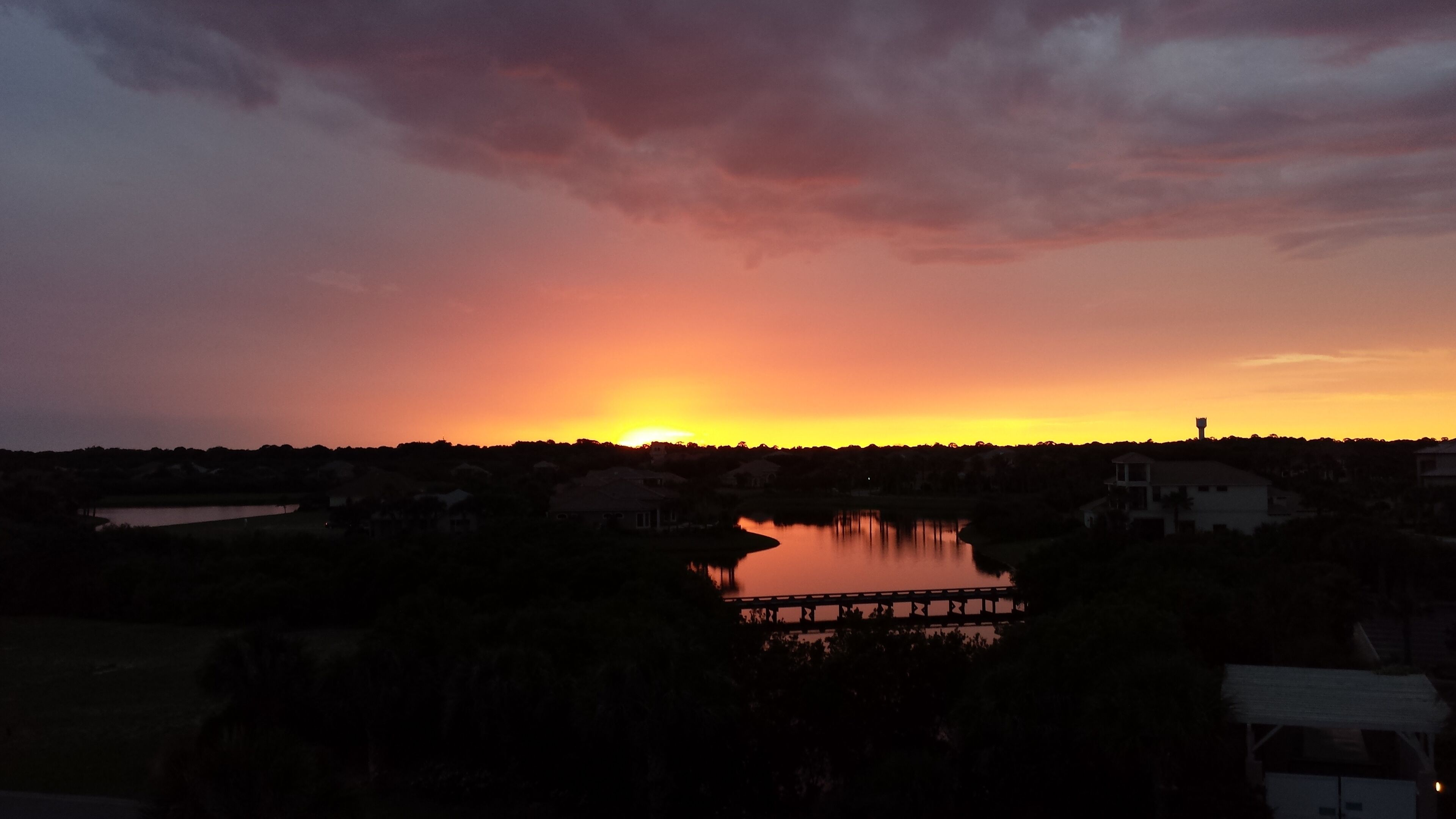 Solnedgang over en sø med en bro og huse i forgrunden.