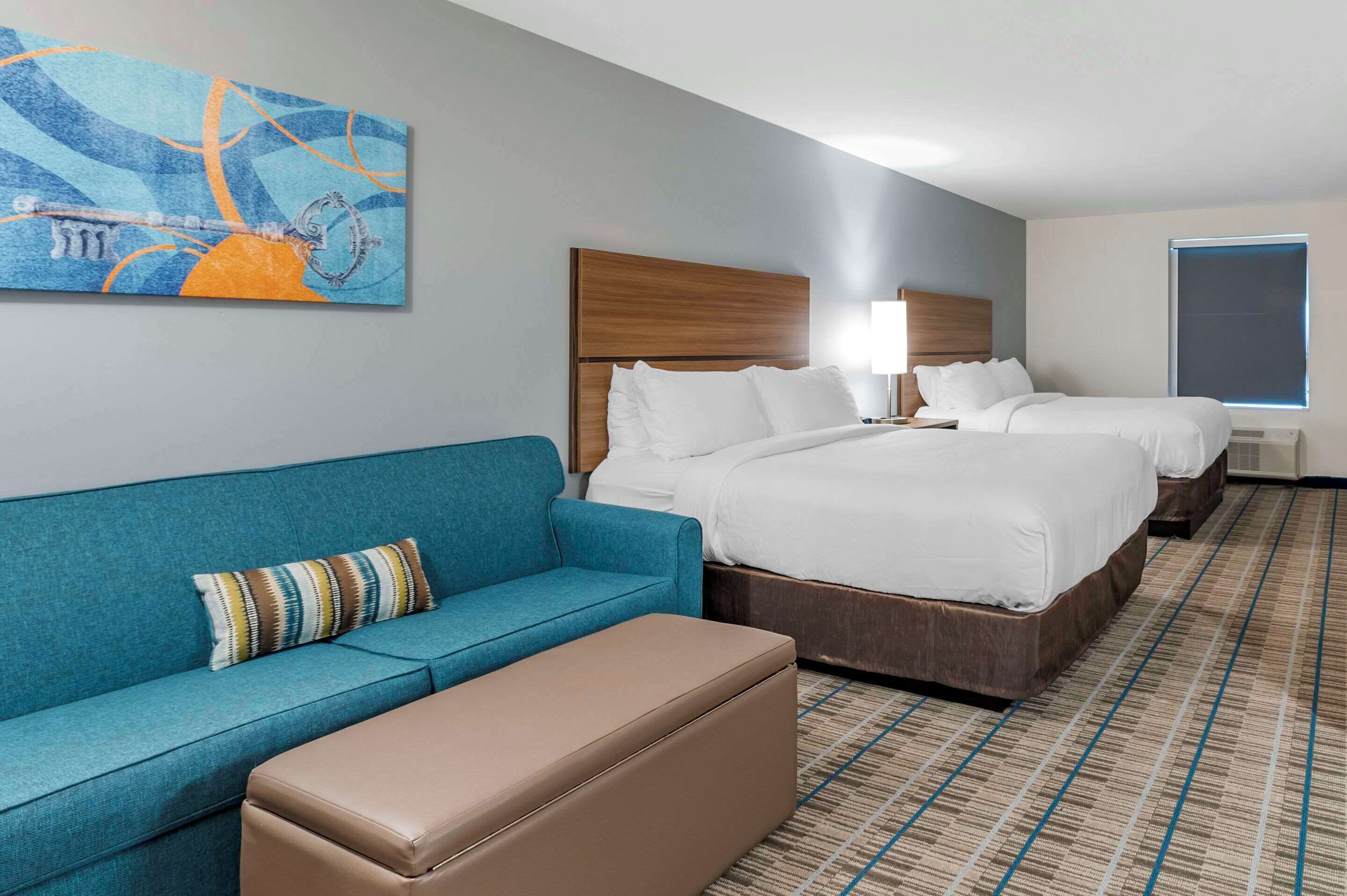 A hotel room with a large bed, a blue sofa, a brown ottoman, and a painting on the wall.
