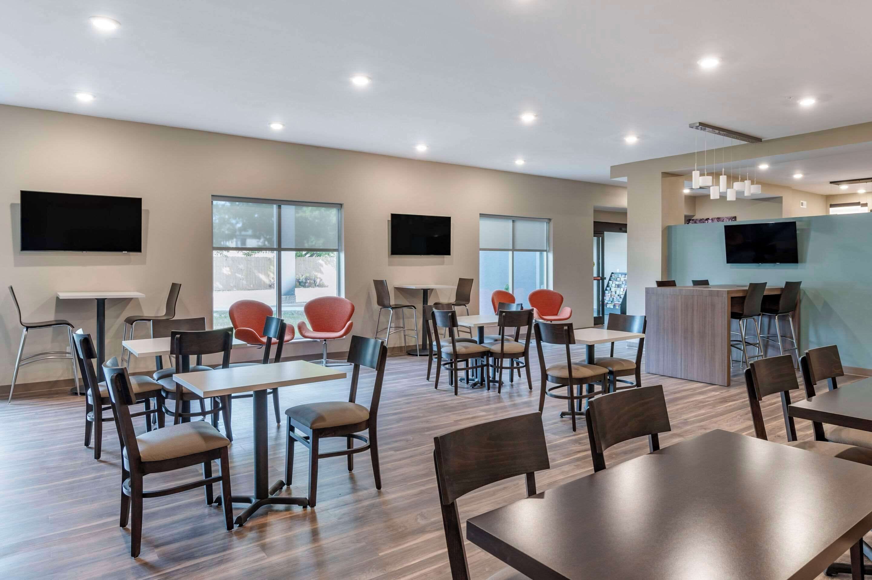 A modern dining area with multiple tables and chairs, large windows, and a flat-screen TV mounted on the wall.