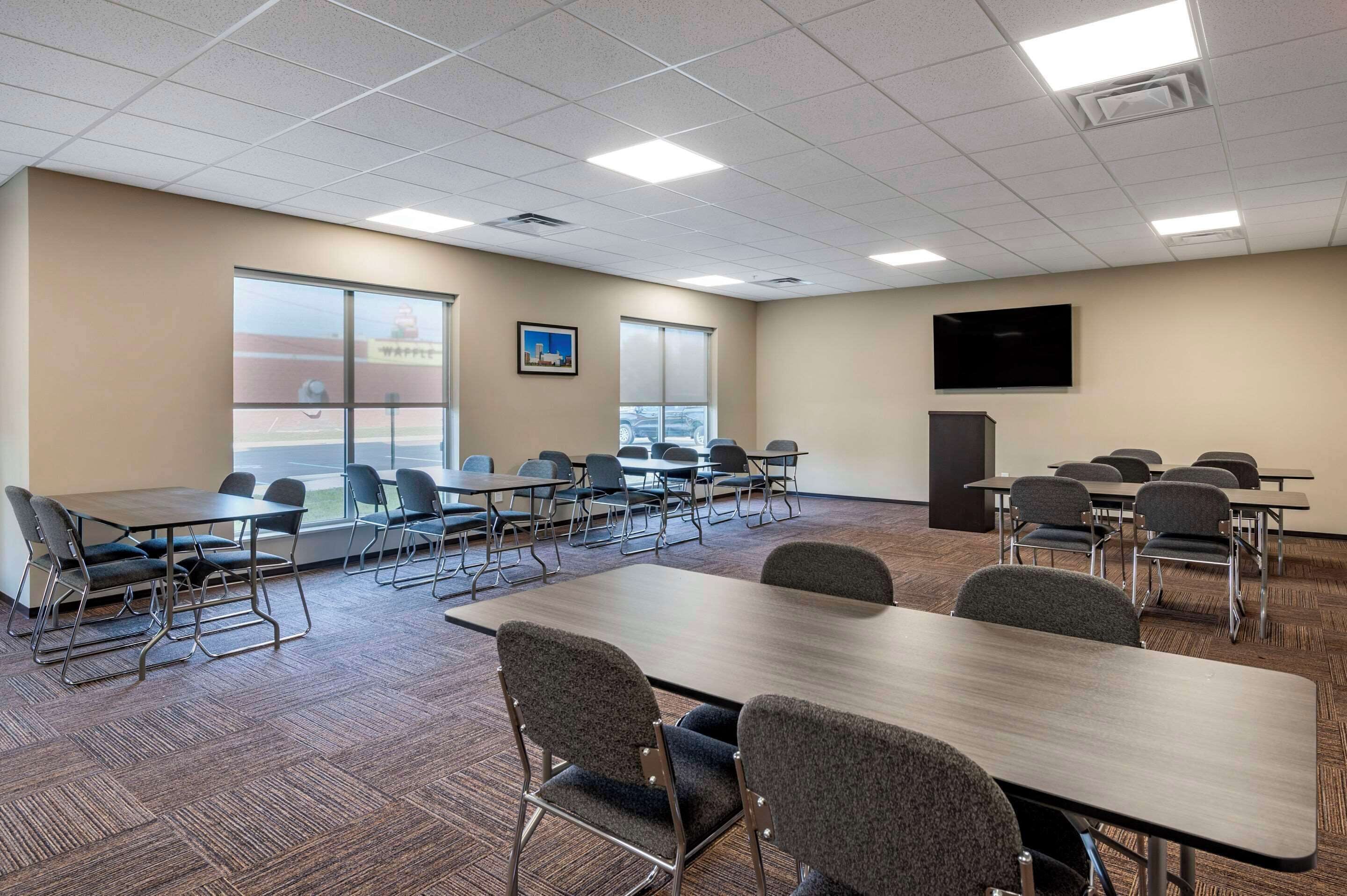 A conference room with multiple tables and chairs, large windows, a flat-screen TV, and a carpeted floor.