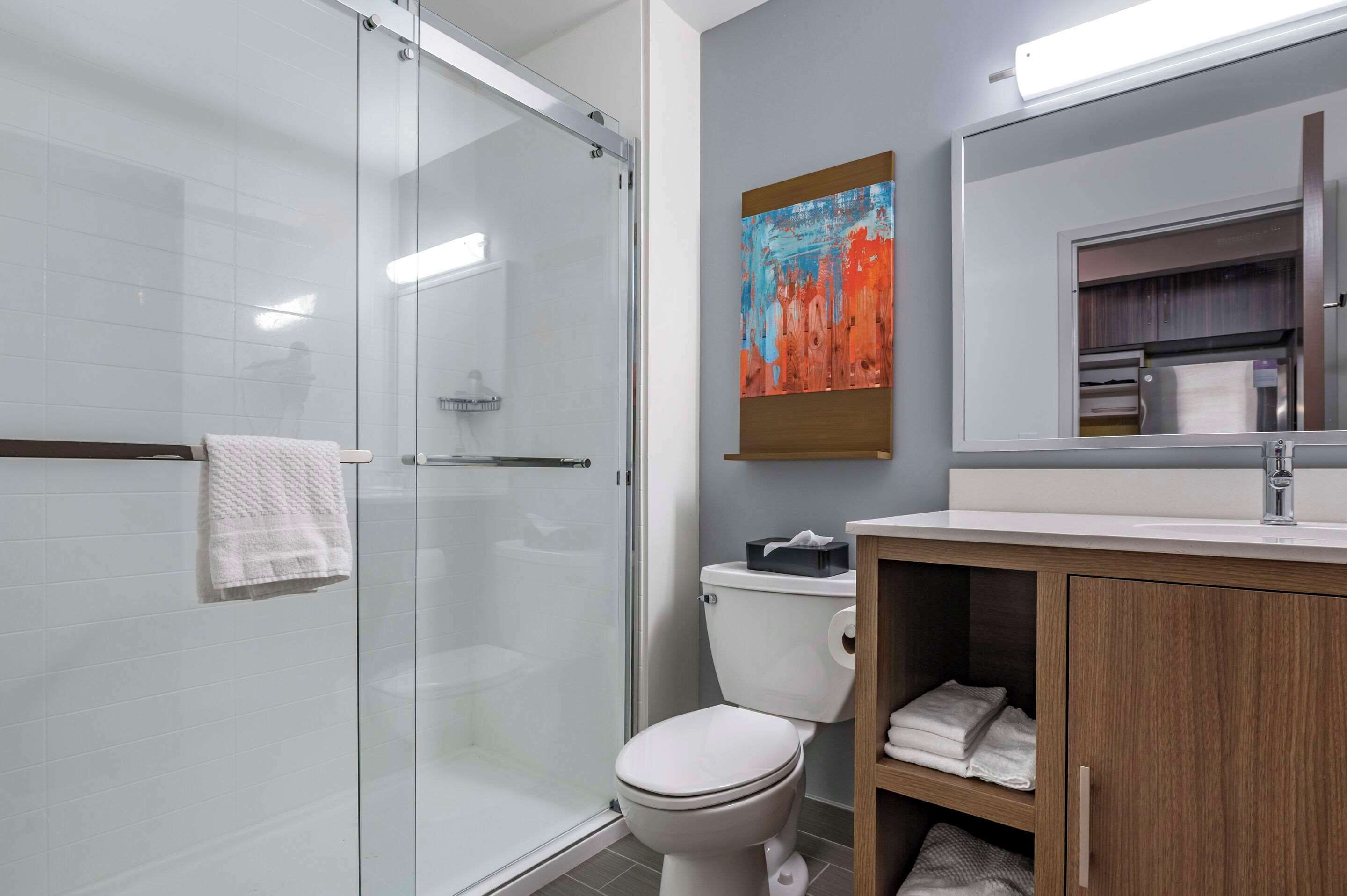 Un baño moderno con cabina de ducha de vidrio, inodoro blanco, tocador de madera con espejo y un cuadro abstracto colorido en la pared.