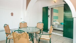 Un comedor con una mesa redonda y cuatro sillas de mimbre, un arco de entrada y una cama con contraventanas verdes al fondo.
