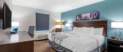 A hotel room with a large bed, a desk with a computer, a flat-screen TV, and a window with blinds.