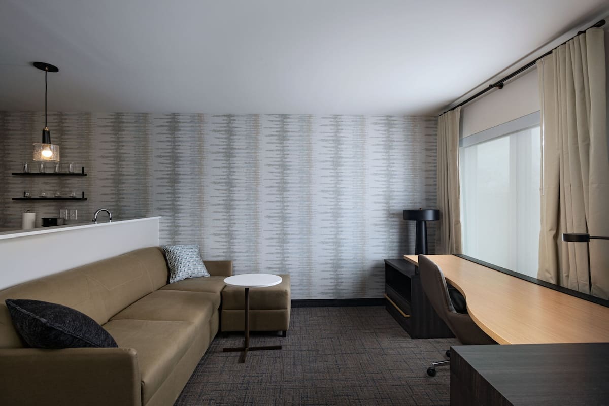 A modern hotel room with a sofa, a small round table, a desk, a TV, a lamp, and a large window with curtains.