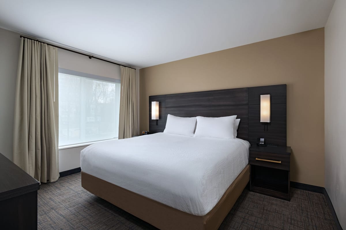 A hotel room with a large bed, two bedside tables, a headboard with wall sconces, and a window with curtains.