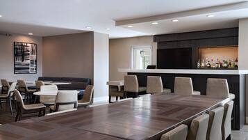Un espace repas moderne avec une longue table en bois et plusieurs chaises, un bar équipé d’un téléviseur et une grande peinture abstraite accrochée au mur.