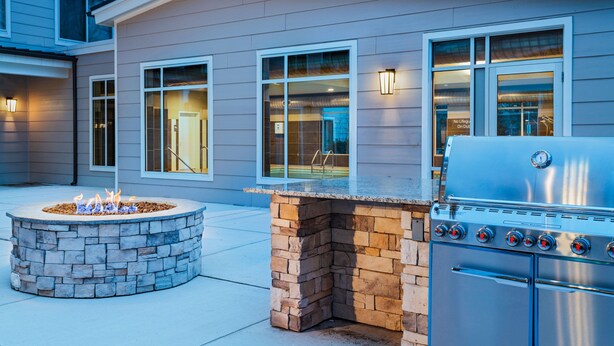 Una cocina exterior moderna con un hogar de piedra y una parrilla de acero inoxidable, ubicada frente a una casa con ventanas amplias e iluminación exterior.