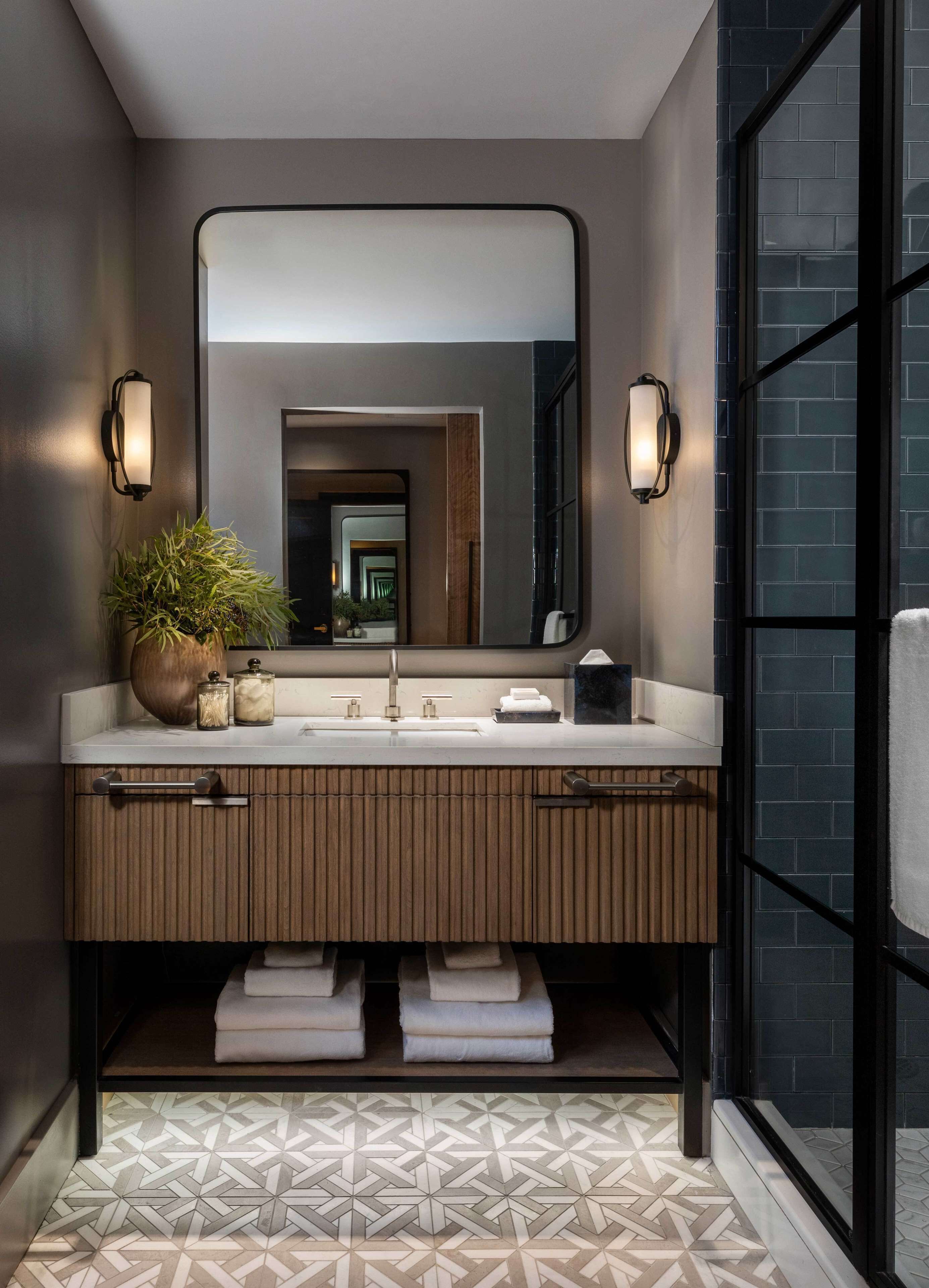 A modern bathroom with a large mirror, two wall-mounted lights, a wooden vanity, and a geometric patterned floor.