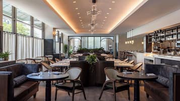 A modern restaurant interior with round tables, cushioned seating, a central bar area, and large windows providing natural light.