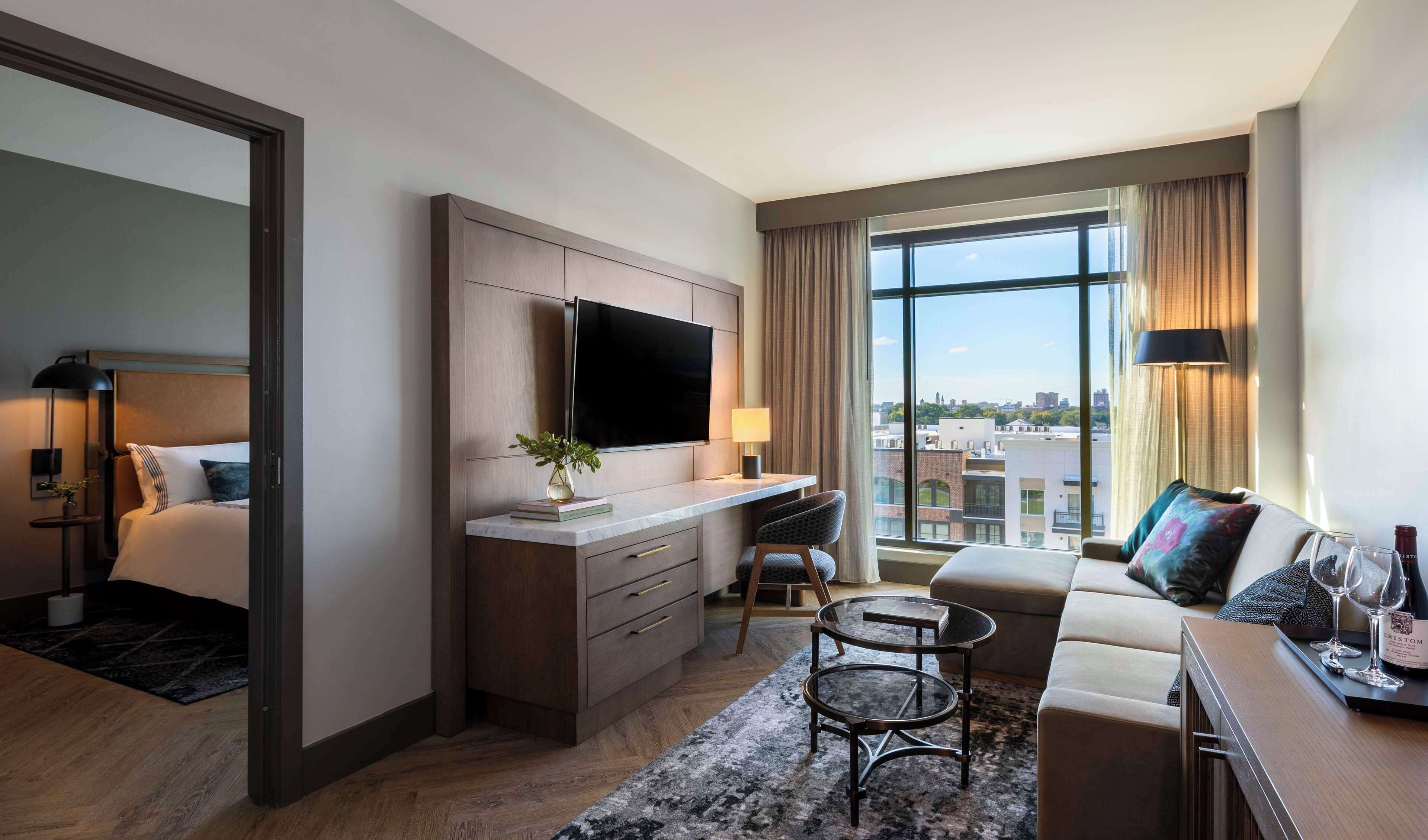 A hotel room with a bed, a desk with a flat-screen TV, a sofa, and a large window offering a city view.