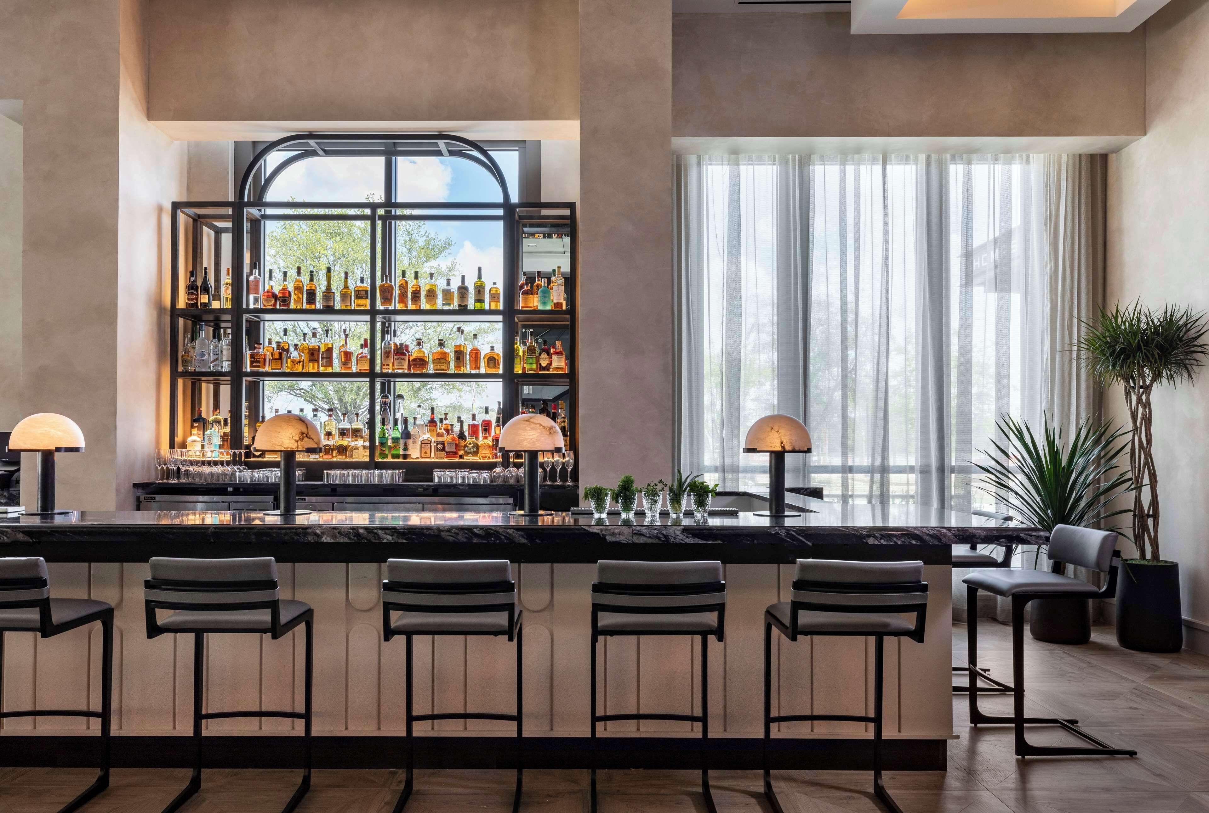 A bar area with a high shelf displaying various bottles, bar stools, and a large window with sheer curtains.