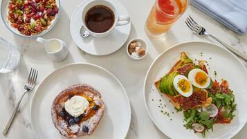 A breakfast setting with various dishes including a fruit salad, coffee, a glass of juice, a plate with eggs and bacon, and a dessert with whipped cream and blueberries.