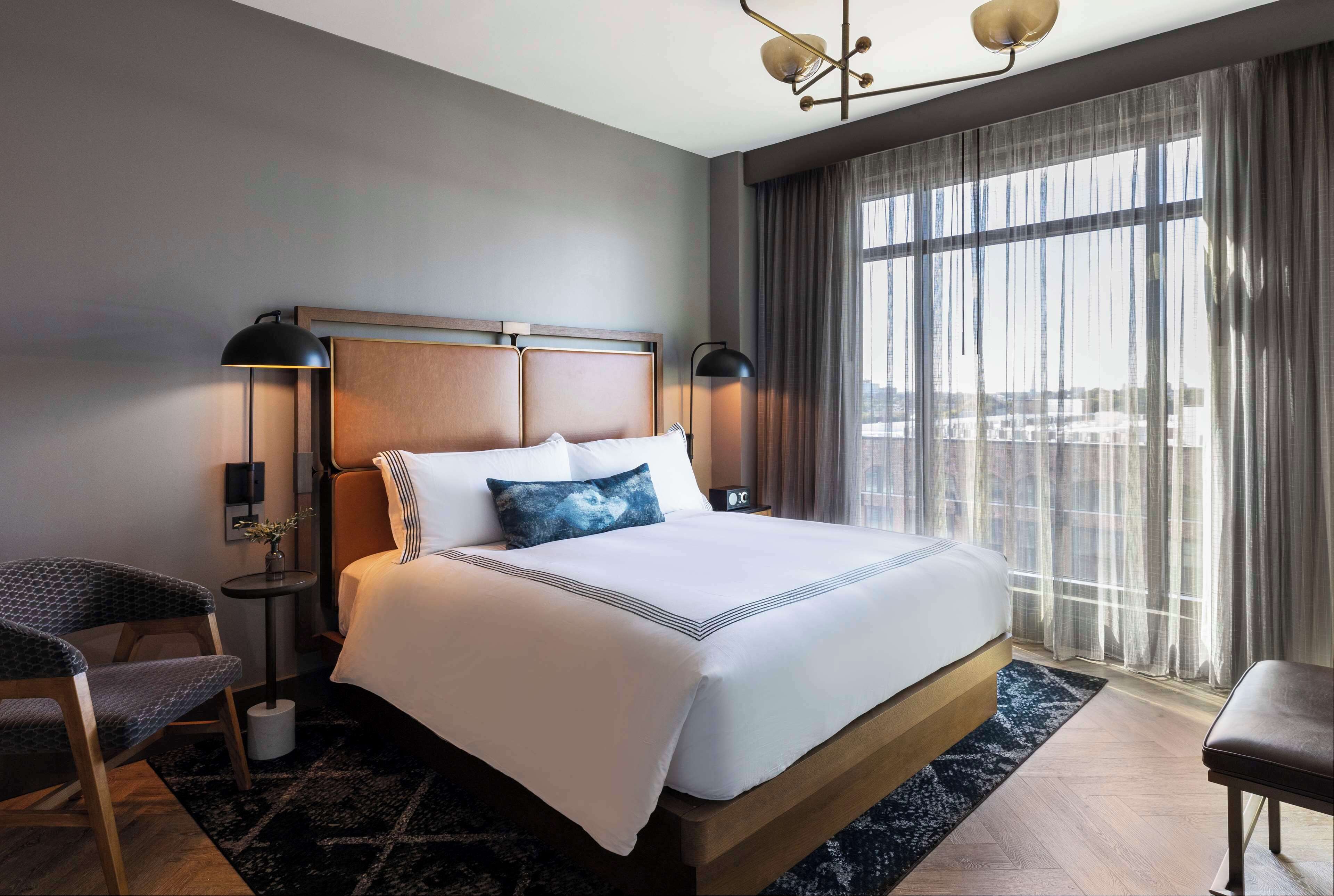 Une chambre d’hôtel avec un grand lit, deux lampes de chevet, une chaise, une petite table et une vue sur la ville à travers de fines rideaux.